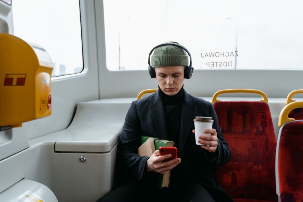 Man on a bus with headphones and a coffee, looking at his phone