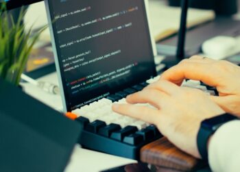 Hands typing on a mechanical keyboard beside a code-filled laptop and green plant for a Tailscale Ubuntu guide.